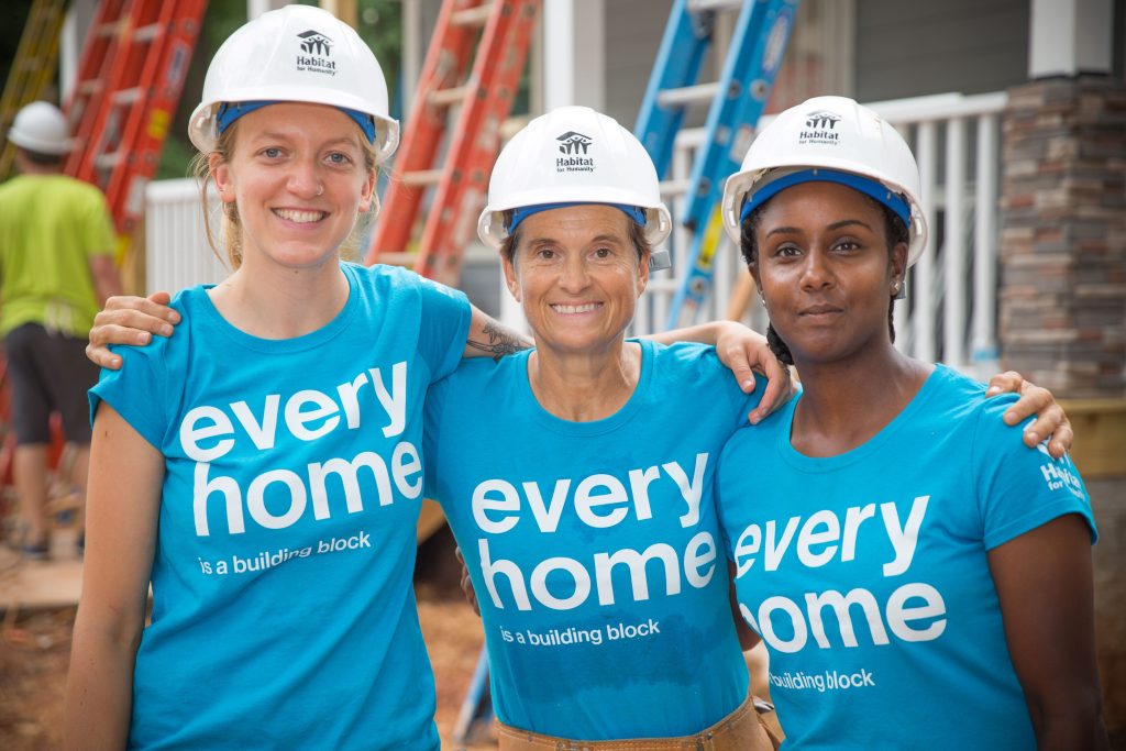 3 women volunteers standing in a group hug outside a Habitat building site.